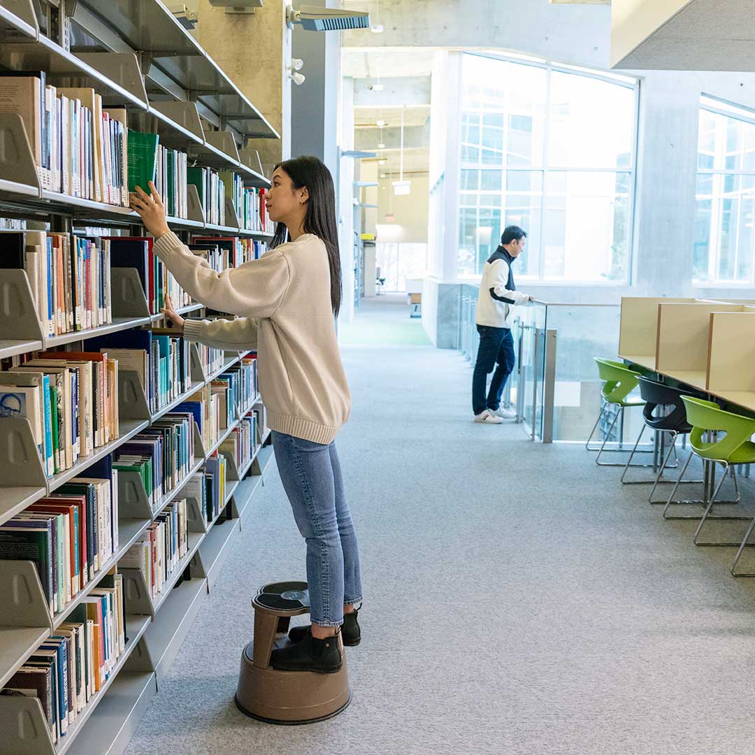 On the third floor of the library, a determined student is on a quest, her eyes scanning the shelves for the perfect book to complete her final assignment.
