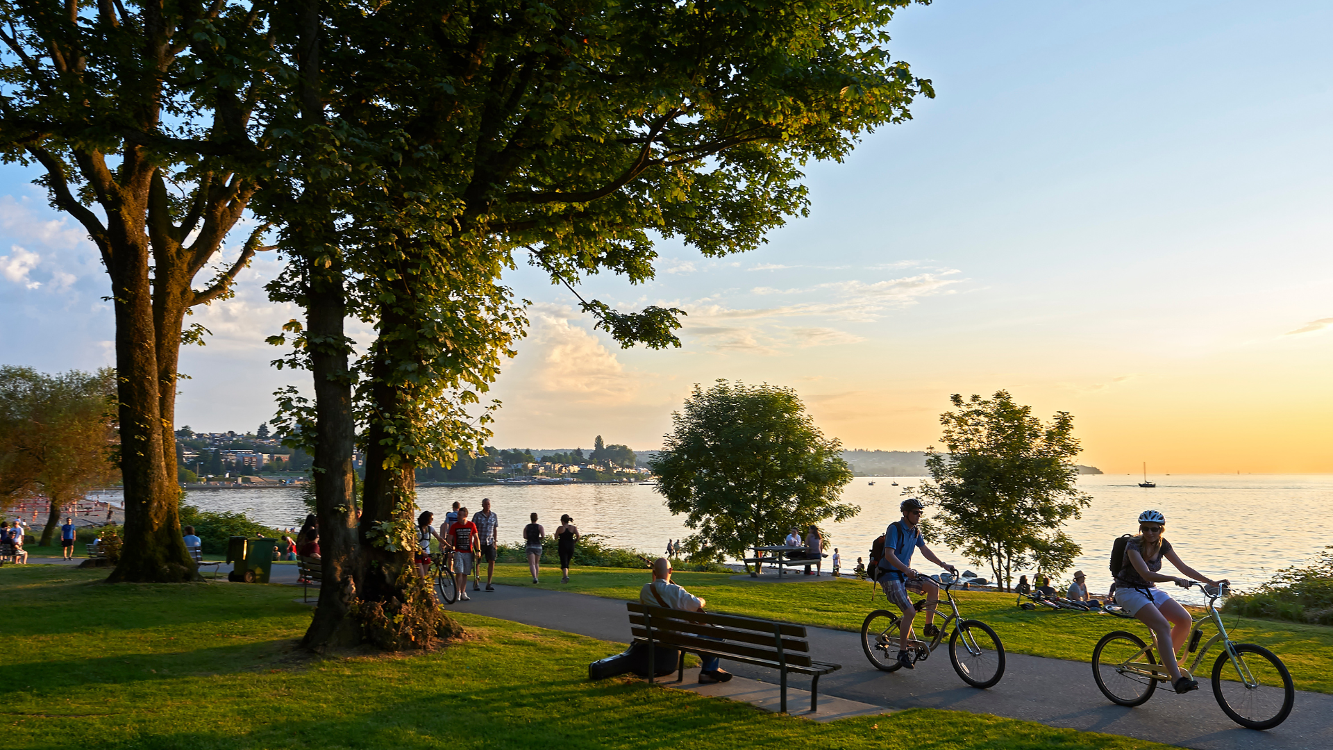 Kitsilano Beach at sunset. A man plays a guitar on a bench while walkers and bikers pass by in front of him.