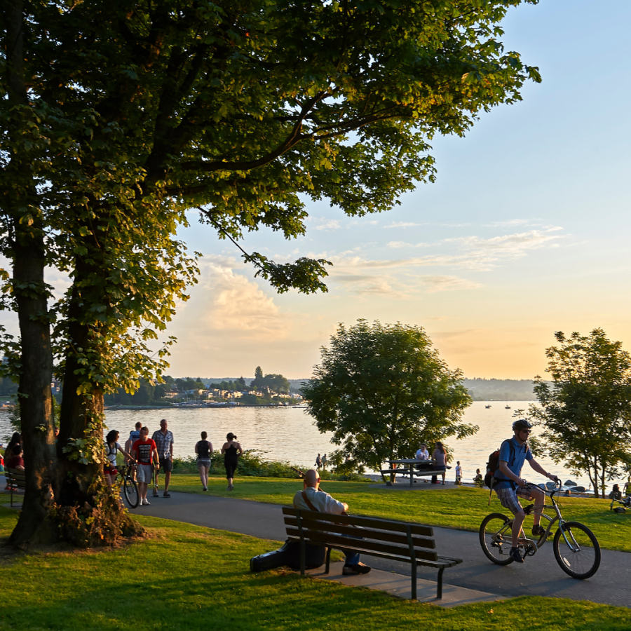 Kitsilano Beach at sunset. A man plays a guitar on a bench while walkers and bikers pass by in front of him.
